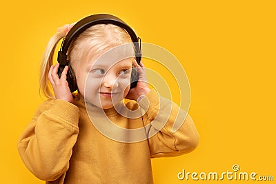 Cute flirtatious fair-haired preschool girl listens to music with headphones. Close up portrait on yellow background Stock Photo