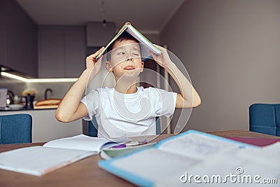 Cute funny small Caucasian teen schoolboy holding book above head and mocking. Lazy,homework. Stock Photo
