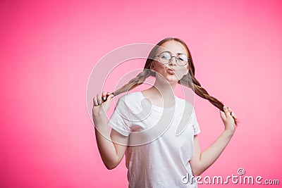 Cute funny young student girl in white t-shirt, glasses and braid shows kiss lips on a pink background with copy space Stock Photo