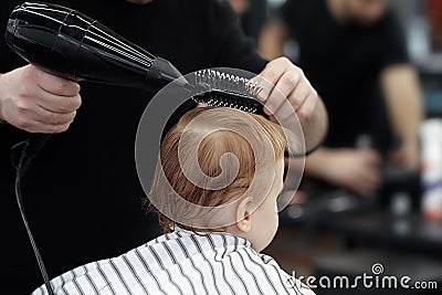 Cute ginger baby boy in a barber shop having haircut by hairdresser. Hands of stylist with tools hair dryer and hairbrush. Stock Photo