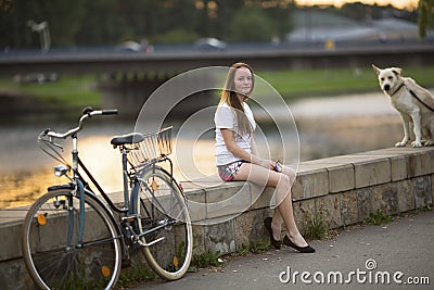 Cute girl with a bicycle and a dog sitting on the promenade in the soft sunset light. Stock Photo