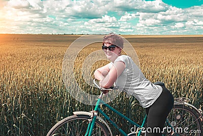 Cute girl with ginger hair standing near the old bicycle. Pretty woman and vintage bike among of golden wheat fields at sunny summ Stock Photo