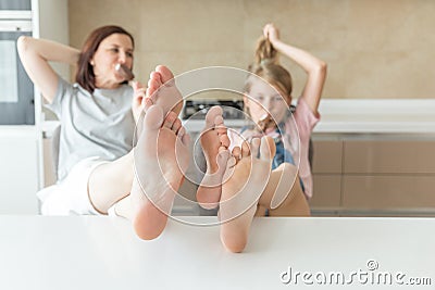 Cute girl and her mother are smiling while eating ice cream in the kitchen with legs on a table Stock Photo