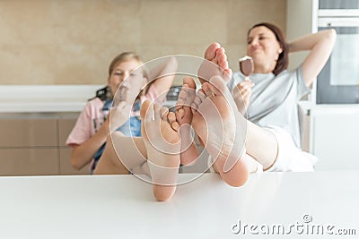 Cute girl and her mother are smiling while eating ice cream in the kitchen with legs on a table Stock Photo
