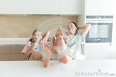 Cute girl and her mother are smiling while eating ice cream in the kitchen with legs on a table Stock Photo