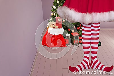 Cute girl with pointed elven feet wearing elf legging standing in a room with a christmas tree and santa claus bag full of Stock Photo