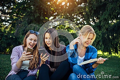 Cute girl students spend free time sitting on green grass in the park on summer day. One woman is reading a book and two young Stock Photo