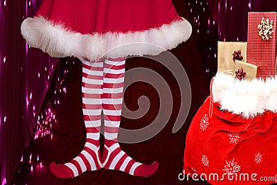 Cute girl wearing red white striped elf stockings and a cute dress standing next to a santa claus bag full of presents on a Stock Photo