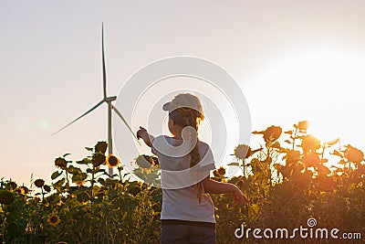 Cute girl in white t-shirt smelling sunflower in sunset field wind turbines farm on background. Child with long braid Stock Photo