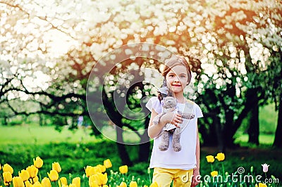 Cute happy child girl holding teddy bear on spring walk with yellow tulips on background Stock Photo