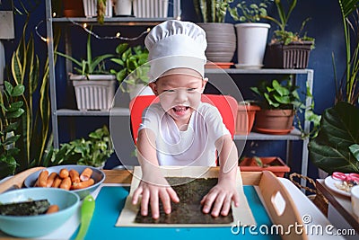 Cute happy smiling Asian little boy child wearing chef hat having fun preparing, cooking healthy Japanese food - sushi roll at Stock Photo