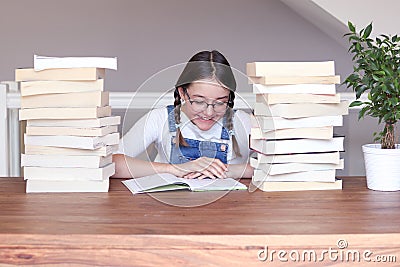 Cute happy smiling tween girl in glasses studying reading book sitting a table with pile of books at home. Stock Photo