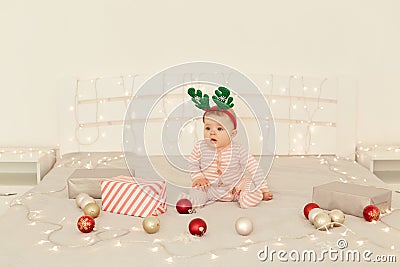 Cute infant girl sitting on Christmas decorations bed and wearing striped long sleeve baby sleeper and festive deer horns, looking Stock Photo