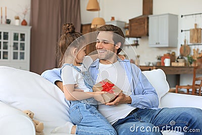 Cute kid daughter make surprise to daddy, little girl presents gift box to father sit on sofa. Father`s day Stock Photo