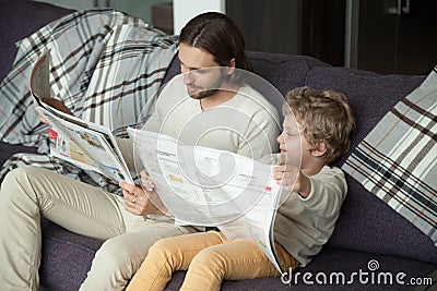 Cute kid son reading newspaper sitting on sofa with dad Stock Photo