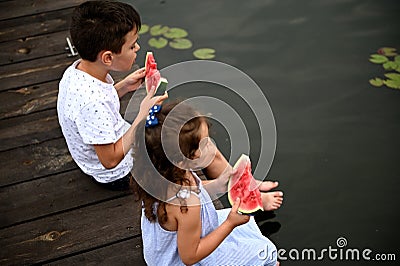 Cute kids with slices of watermelon in their hands, sitting on the pier with their legs lowered into the water and enjoying summer Stock Photo