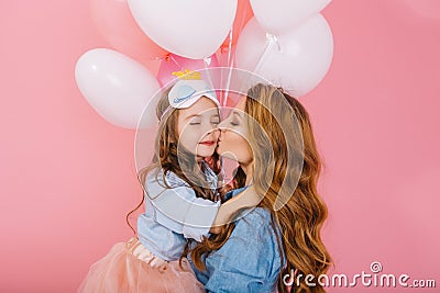 Cute kiss of young curly mom in denim shirt and lovely daughter in sleep mask at birthday party. Little long-haired girl Stock Photo