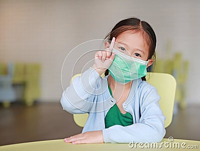 Cute little Asian child girl wearing a protective mask with showing one forefinger sitting on kid chair in children room Stock Photo