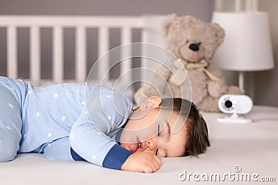 Cute little baby boy in light blue pajamas sleeping peacefully on bed at home with baby monitor camera and soft teddy bear toy at Stock Photo