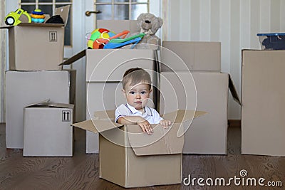 Cute little baby boy sitting inside cardboard box with big boxes full of toys on background, moving out Stock Photo