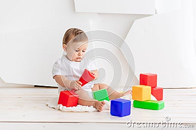Cute little baby six months old in a white t shirt and diapers playing at home on a Mat in a bright room with bright colored cubes Stock Photo