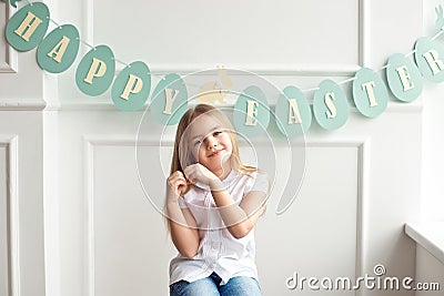 Cute little blond girl sitting in a bright room near the window on the background of streamers with the inscription Stock Photo