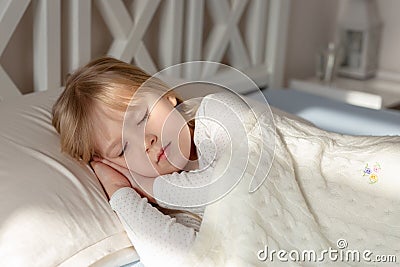 Cute little blond toddler girl sleeping in bed.Sweet baby lying with closed eyes under rays of sun at sunrise in early Stock Photo