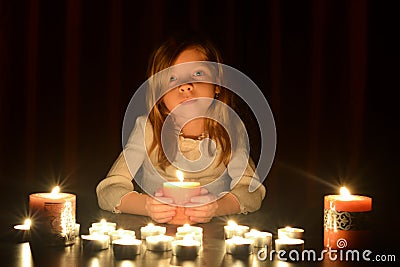 The cute little blonde girl is holding a burning candle, lots of candles are around her over dark background. Stock Photo
