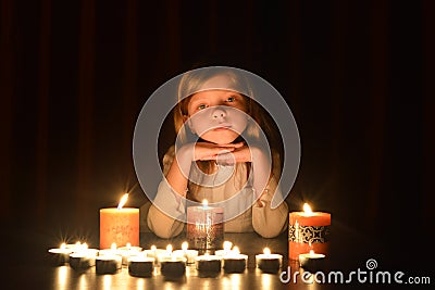 The cute little blonde girl keeps her hands under the chin. Lots of candles are around her, over dark background Stock Photo