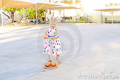 Cute little blondy toddler girl in dress riding scooter in the city park recreation area with canopy, umbrellas. Active leisure in Stock Photo