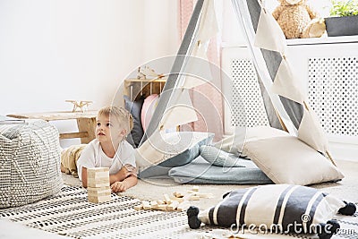 Cute little boy lying on the floor of scandinavian playroom with grey tent with cozy pillows and patterned carpet, real photo with Stock Photo