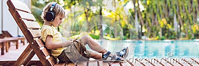 A cute little boy is using a smartphone and headphones lying on a deckchair by the pool. primary education, friendship, childhood, Stock Photo