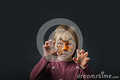 Cute little caucasian girl with tiger face painting on the black background. Close up portrait of little kid with face-painting. Stock Photo