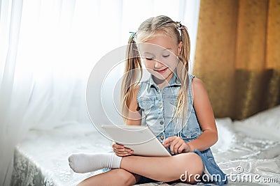 Cute little child girl blond in denim sundress lies in bed uses digital tablet. child playing on tablet pc having fun and smiles Stock Photo