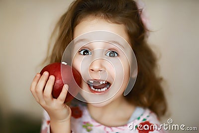 A cute little curly toothless girl smiles and holds a red apple, a portrait of a happy baby eating a red apple, the child loses Stock Photo