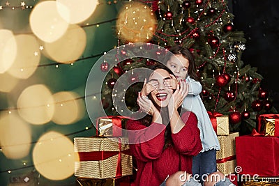 Cute little girl covering her mother eyes. Beautiful room with christmas tree and holiday decoration Stock Photo