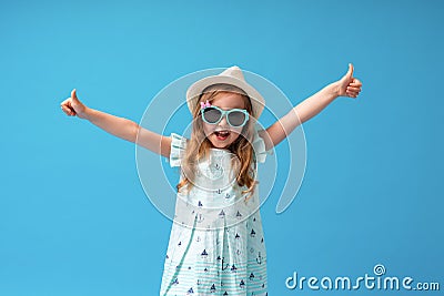 Cute little girl in a dress, hat and sunglasses poses on a blue background Stock Photo