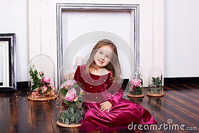A cute little girl in a dress is sitting on the floor with a rose in a flask. Looking at the camera. Childhood. Sweet princess. Th Stock Photo