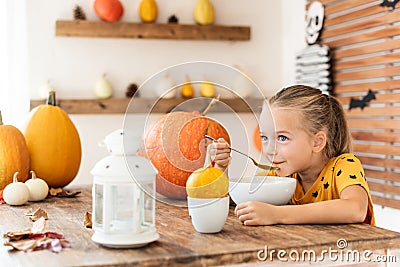 Cute little girl eating pumpkin soup in Halloween decorated dinning room. Autumn season comfort food. Stock Photo