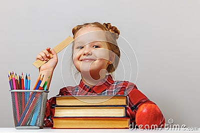 A cute little girl with glasses is sitting at the table, holding a ruler in her hand. Schoolgirl is surrounded by books. Stock Photo