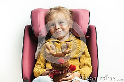 Cute little girl holding stuffed Rudolph Red-Nosed Reindeer toy while sitting in a safety car seat. Kid safety while traveling by Stock Photo