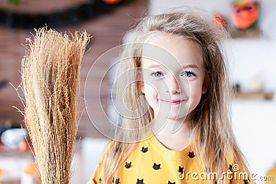 Cute little girl with messy hair, dressed up as a witch and holding a broom is standing in Halloween decorated living room,smiling Stock Photo