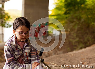 Cute little girl in a plaid shirt and glasses sits sadly under a bridge Stock Photo