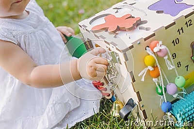 Cute little girl is playing with busiboard outdoors on green grass. Educational toy for toddlers. girl opened door to cube of Stock Photo