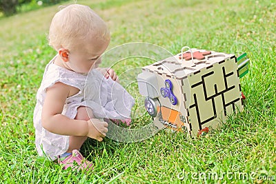 Cute little girl is playing with busiboard outdoors on green grass. Educational toy for toddlers. girl opened door to cube of Stock Photo