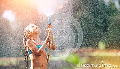 Cute little girl pours herself from the hose, makes a rain. Hot summer day pleasure Stock Photo