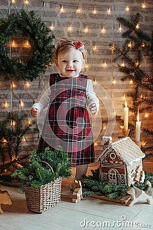 Cute little girl is sitting by the Christmas tree on the floor in the room. Near the baby is a gingerbread house and a basket with Stock Photo