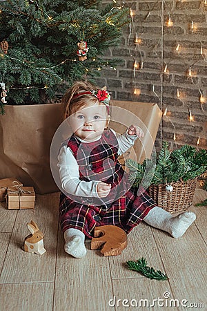 Cute little girl is sitting by the Christmas tree on the floor in the room. Near the baby is a gingerbread house and a basket with Stock Photo