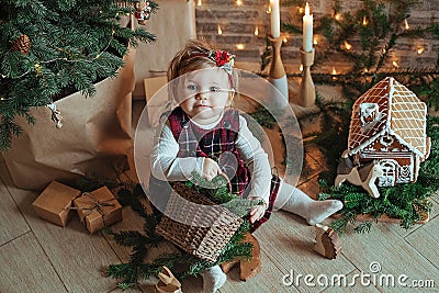 Cute little girl is sitting by the Christmas tree on the floor in the room. Near the baby is a gingerbread house and a basket with Stock Photo