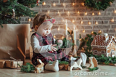 Cute little girl is sitting by the Christmas tree on the floor in the room. Near the baby is a gingerbread house and a basket with Stock Photo